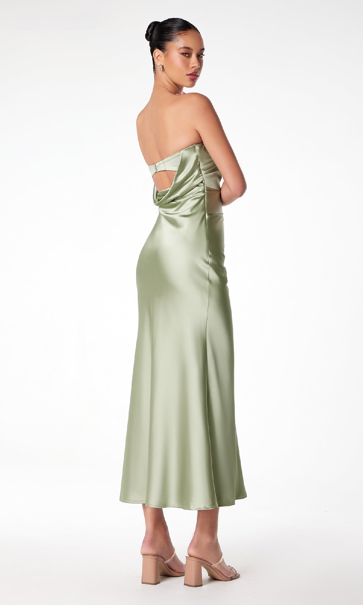 A woman in the Strapless Long Satin Maxi Prom Dress: Astee T2006, light green with an open back and strappy detail, stands facing away and looks over her shoulder. She pairs it with nude heels and a bun hairstyle.